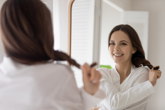Happy young woman in bathrobe pulling tuft of long, smooth string hair, looking in mirror, smiling at reflection. Consumer satisfied with haircare cosmetic product effect, shampoo, balm, conditioner.