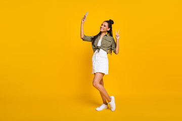 Photo portrait full body view of dark skin woman showing v-sign taking selfie with phone isolated on vivid yellow colored background