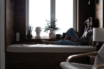 Fototapeta premium Beautiful young woman listening to music while sitting on sofa at home near the window