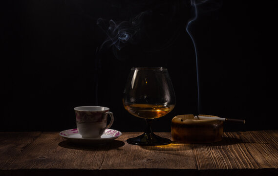 Background image of a wooden bar in a pub with a glass of cognac and a smoking cigarette in an ashtray and a cup of coffee on a dark background with a bokeh effect