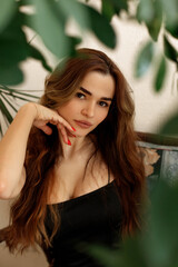 Fototapeta premium Beautiful women in greenery looking at the camera, sitting on the chair on terrace at home. Portrait of a girl with long hair brown-haired, tropical ethno boho style interior