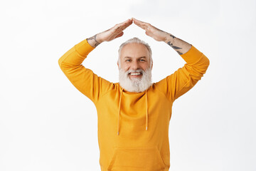 Happy senior guy with tattoos make roof with hands above head, showing family gesture, home rooftop and smiling, wearing orange hoodie, standing over white background