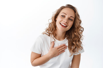 Grateful blond girl smiling, holding hand on heart and looking aside at promotional text with thankful and pleased face, standing over white background