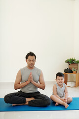 Laughing little boy sitting next to his father trying to meditate at home