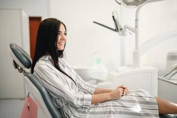 Smiling and satisfied patient in a dental office after treatment. Happy woman patient sitting on dentist chair in dental clinic. Dentistry care concept.