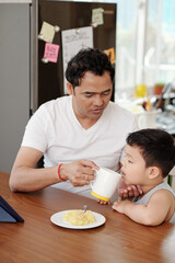 Father giving his little son a cup of water when they are eating breakfast and watching animated...