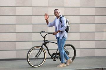 Positive millennial guy with modern bike waving near brick wall outdoors