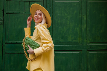 Joyful smiling woman wearing yellow hat, classic shirt, holding quilted faux leather green bag with...
