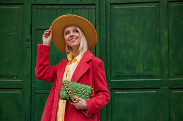 Fototapeta premium Happy smiling fashionable woman wearing stylish outfit with yellow hat, coral color coat, holding quilted faux leather bag, posing on green background. Model looking up. Copy, empty space for text