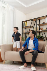 Smiling Indian man looking at his excited little son jumping on sofa and singing
