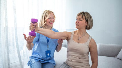 Fototapeta premium Health visitor and a mature woman during home visit. A nurse or a physiotherapist helping a smature woman exercise with dumbbells. A Modern rehabilitation physiotherapy in the room
