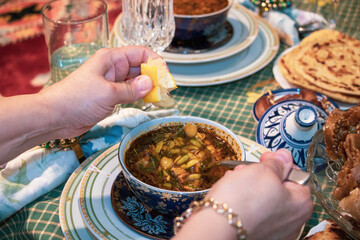 TWO HANDS OF AN ADULT WEARING A RING AND BRACELET ON A BOWL OF HEALTHY VEGETABLE SOUP, ONE HAND PUTS LEMON AND THE OTHER HANDS A SPOON IN THE BACKGROUND OF OTHER FOOD DISHES, close up