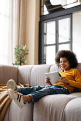 Black curly boy smiling and using tablet computer while sitting on sofa