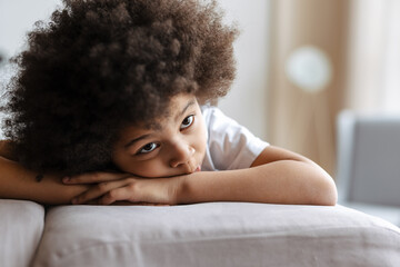 Black curly boy looking aside while lying on sofa at home