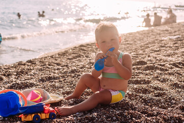 little girl in a swimsuit sits by the sea and plays with sand toys in the summer in the contour light of the sun