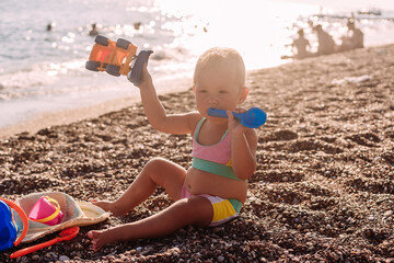 little girl in a swimsuit sits by the sea and plays with sand toys in the summer in the contour light of the sun