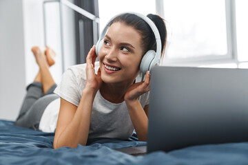 European smiling woman in headphones using laptop while lying on bed