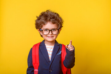 Child boy with book and backpack isolated on yellow paper wall. Happy smiling kid go back to...