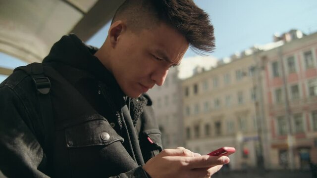 A young Asian man stands at a bus stop and looks into a mobile phone. Tracking city transport using a mobile device. City application. Modern infrastructure. Electro tram or bus arrived on schedule. 