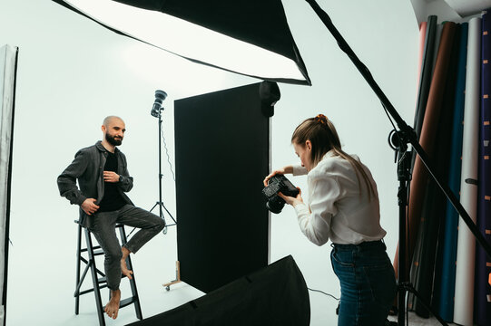 Woman photographer makes a photo shoot for a positive bearded man in a photo studio on a white background. Backstage