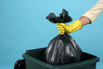 Woman throwing garbage bag into bin on light blue background, closeup