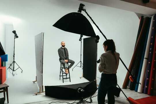 Woman photographer makes portraits for a handsome young man, sits in a chair on a white background and poses with a smile on his face.