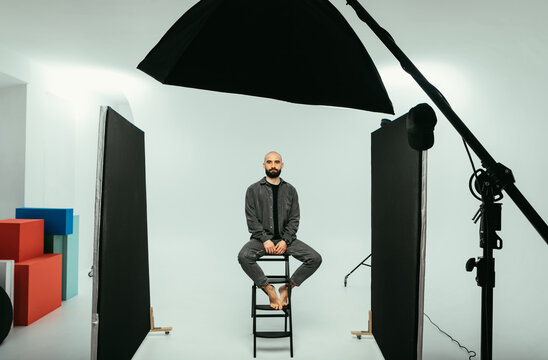 Bearded male model sitting on a chair in a photo studio on a white background and looking at the camera with a serious face.