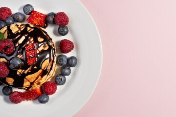 top view of a dessert of pancakes with fruits and chocolate on white plate on pink background. copy space