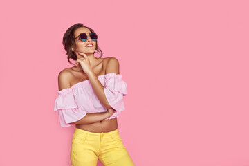 Beautiful girl in yellow jeans and pink shirt, with hands up wearing sunglasses posing, dancing,...