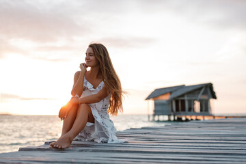 Woman relax at maldives beach, sit on wooden pier end enjoy sunset. With Bungalow in background