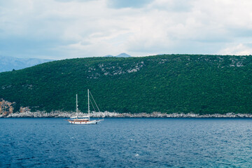 Fototapeta na wymiar White two-masted schooner sails along the rocky coast against the backdrop of mountains