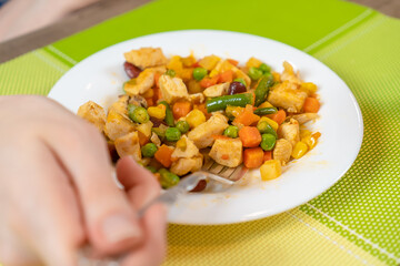 Women's hands eat with a fork fried pieces of meat and a variety of vegetables for lunch, next to a glass of juice. Concept of eating