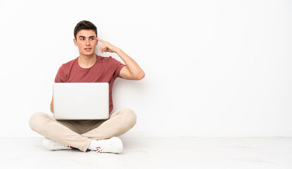 Teenager man sitting on the flor with his laptop making the gesture of madness putting finger on the head