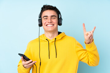 Teenager caucasian man isolated on purple background listening music with a mobile making rock gesture