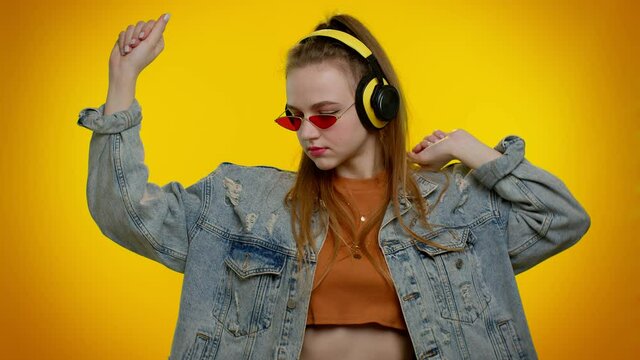 Portrait of teenager girl 20s years old in denim jacket listening music via headphones and dancing disco fooling around having fun expressive gesticulating hands. Young woman on yellow wall background