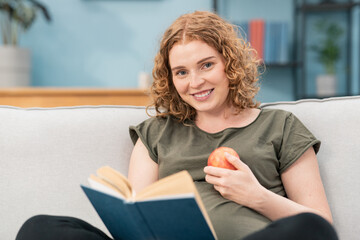 Smiling pregnant woman sitting on sofa reading book and holding apple in one hand.