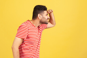 Profile portrait curious bearded man in red striped t-shirt trying to look far away holding hand on forehead, eye vision problems. Indoor studio shot isolated on yellow background