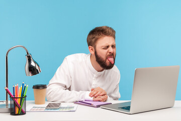 Funny displeased man with beard showing tongue out talking video call on laptop sitting at...