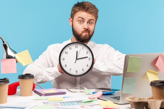 Funny unhappy man with beard in white shirt showing big wall clock sitting at workplace all covered with sticky notes, in hurry to finish his work. Indoor studio shot isolated on blue background