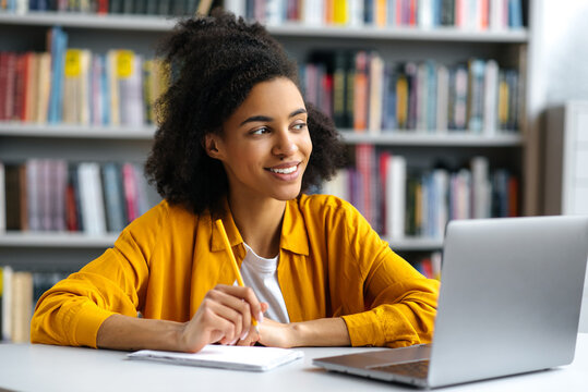 Happy successful pretty curly haired african american female student sitting at table in campus library in casual clothes, with laptop, doing homework or preparing for exam, looking away, smiling
