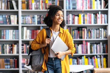 Portrait of joyful pretty african american female student standing against background of bookshelves in university library holding laptop and backpack looking to the side, smiling pleasant - obrazy, fototapety, plakaty