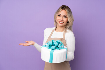 Young pastry chef holding a big cake over isolated purple background extending hands to the side for inviting to come