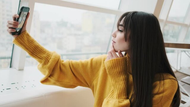 The side view of a woman in a yellow sweater taking selfie on a phone and posing with gestures while sitting in front of a window