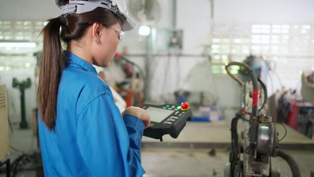 Robotic arm machine was controlled  by engineer woman in the blue uniform standing in the factory. Wearing safety helmet and glasses. Industrial, technology and innovation lifestyle for ai concept