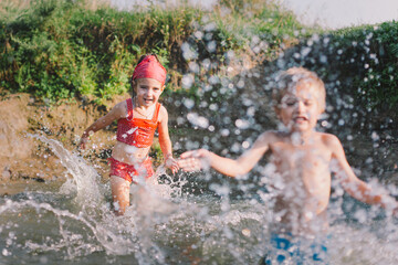 Happy summertime, healthy childhood concept. Caucasian children playing, splashing, jumping and having fun in a river in summer. Selective focus on one kid. Horizontal shot.