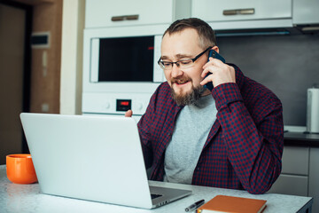 Portrait of stylish intelligent man with beard in modern kitchen interior. Businessman in shirt works at home with a laptop during the quarantine period. Communication with friends in self-isolation.