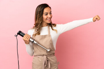 Young woman using hand blender over isolated pink background giving a thumbs up gesture