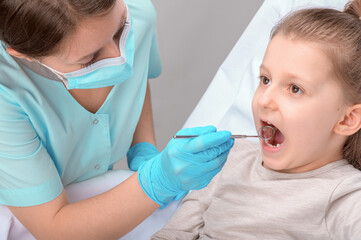 A dentist in a blue uniform performs an examination of the oral cavity and the condition of the teeth of a little girl with the help of a stamotological mirror.