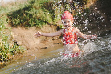 Happy summertime, healthy childhood concept. Little girl playing, splashing, jumping and having fun in a river in summer. Blured green background. Horizontal shot.