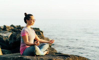 Young woman sitting on a stone pier by the sea meditates in the lotus position. Brunette girl doing yoga workout by the ocean. Healthy sports lifestyle. Mental and physical wellbeing.